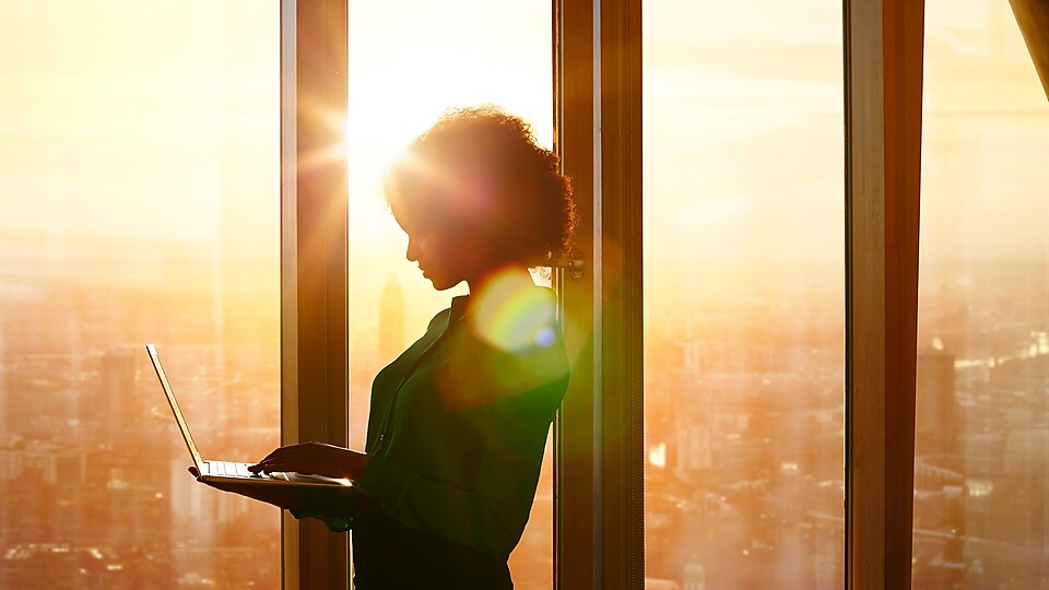 Bir binada pencerelerden görünen gün batımına karşı ayakta dizüstü bilgisayarını kullanan kadın