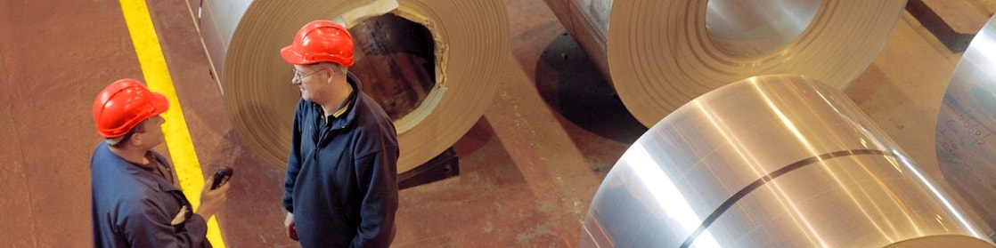 Two factory workers standing next to rolls of steel