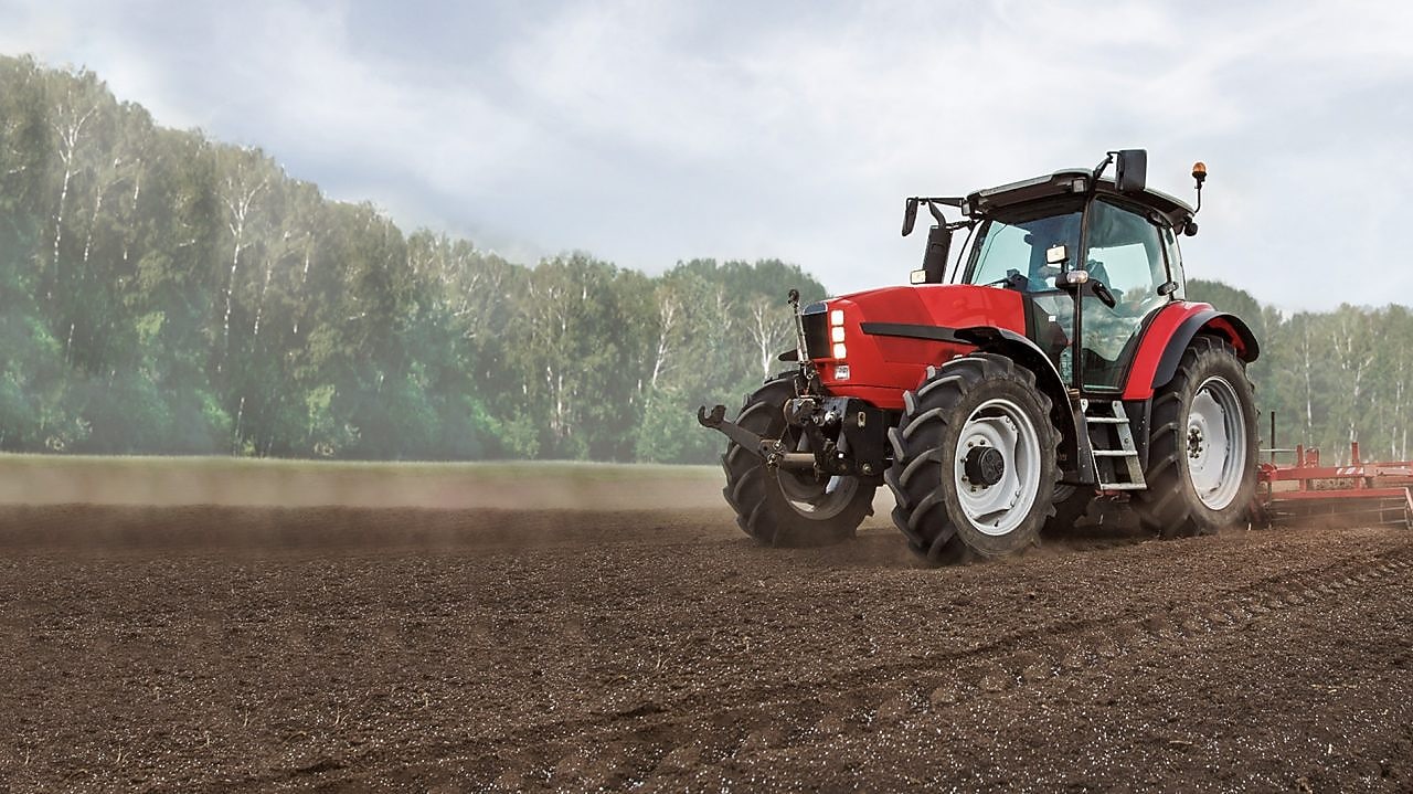 Arka planda ağaçlar bulunan çamurlu bir tarlayı süren kırmızı traktör