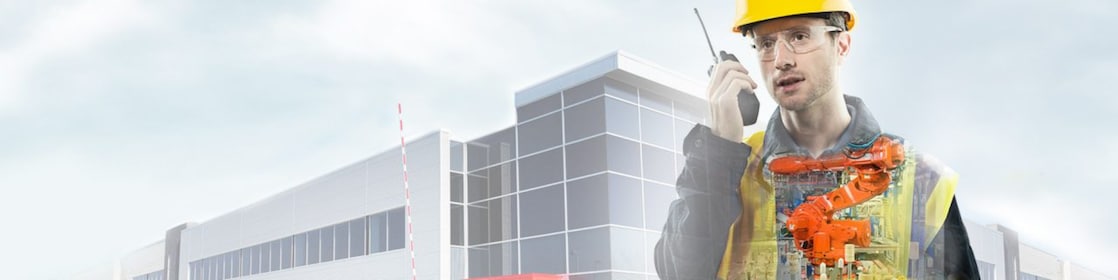 büyük bir binayı gösteren arka planın önünde sarı baretli adam telsizle konuşuyor