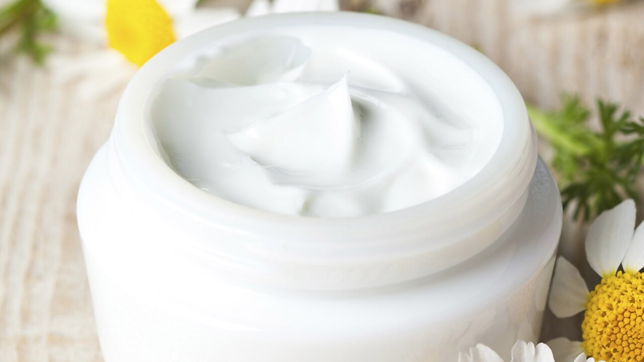 Masa üzerinde çiçeklerle çevrili beyaz bir yüz kremi tüpü