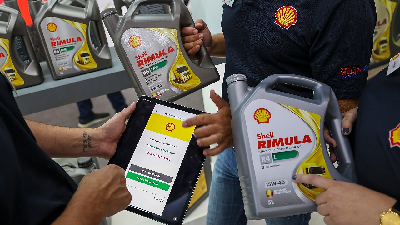 Elinde Shell Rimula ürünleri ve Shell CO2 ve Energy Tracker uygulamarını gösteren bir tablet tutan üç kişi. Arka planda daha fazla Shell Rimula ürünü bulunan raflar gösterilmektedir.